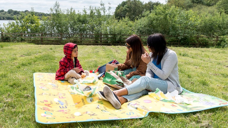 Image shows a family of three sat on a blanket in the grass enjoying a picnic, with views of the water behind them
